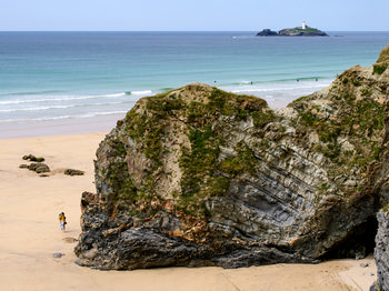 Godrevy Hayle This landscape photograph captures a view of Godrevy at Hayle in Cornwall during the afternoon in early summer. The main focus of the image is the broad sandy beach, with prominent dark rocks and rugged cliffs in the foreground that are dotted with patches of grass and wildflowers. The sea stretches out in gentle waves toward the horizon, where the well-known Godrevy Lighthouse stands on an island just offshore. The clarity of the light suggests mild summer weather, and a few people can be seen walking along the beach, emphasizing the scale of the natural features. The scene combines elements of Cornwall’s coastal landscape, including the cliffs, rocks, sea, and the distinctive lighthouse that marks Godrevy.